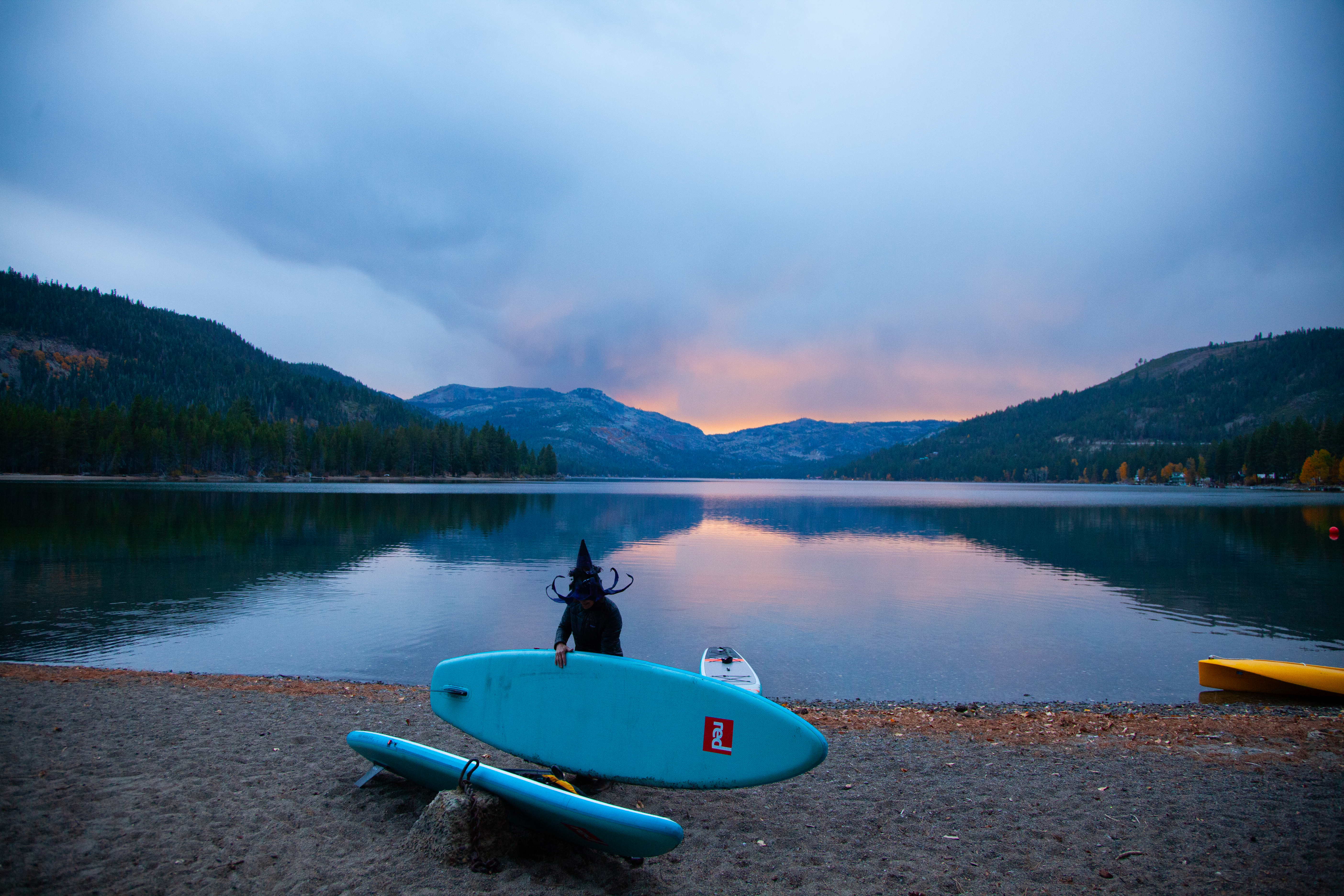 A costumed paddler in a pointed witch hat beside a turquoise paddleboard on the shore of Donner Lake at sunset.